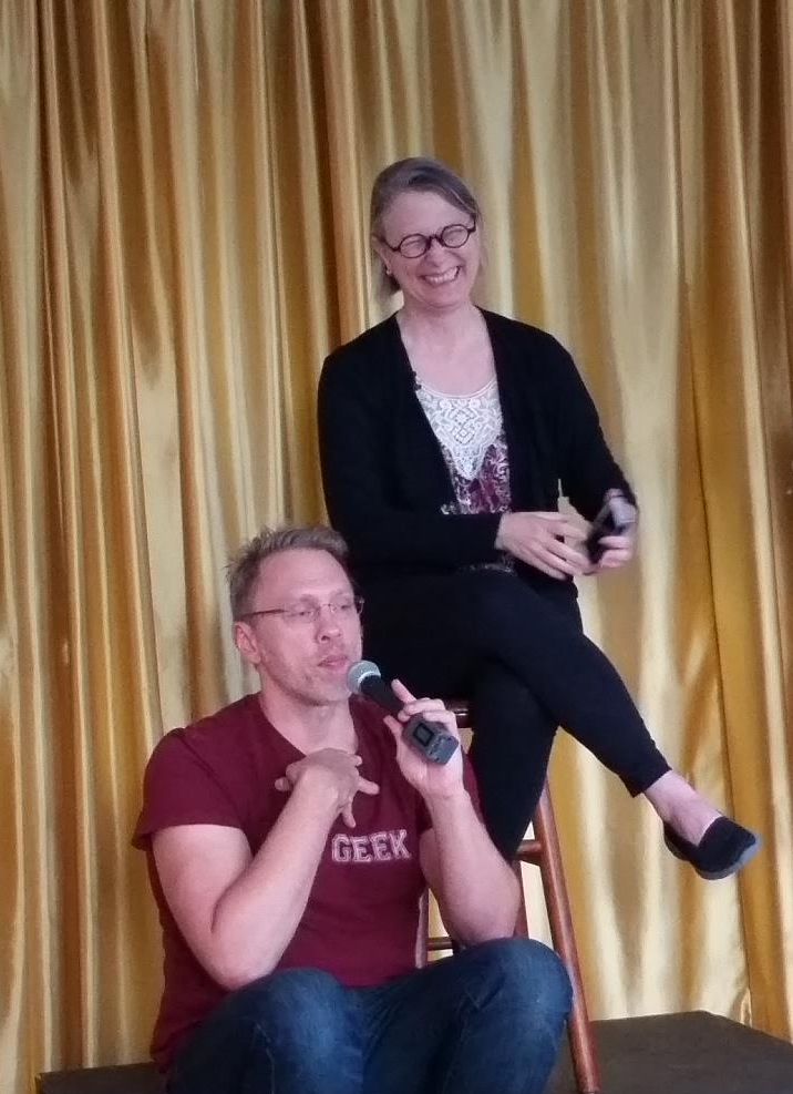 Reid Mihalko and Dr Carol Queen teaching together - Reid —mid-50s white, cis man, wearing a burgundy sex geek t-shirt, with blonde hair and blonde stubble— sitting on a low stage talking on a mic while Carol Queen — white cis woman with pepper hair and artsy rimmed eyeglasses— sits behind and above Reid on a stool laughing