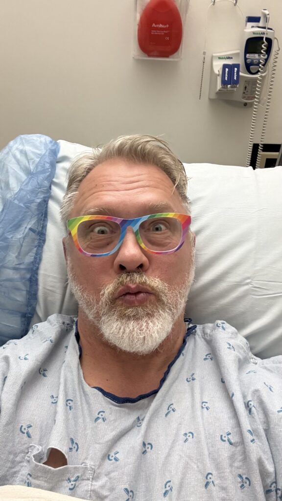 Selfie taken from a hospital bed of Reid, white man in his 50's with grey/blond beard, wearing a light blue/grey hospital gown