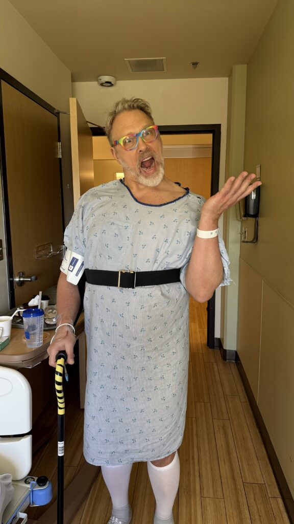 Reid -50+ year old white, cis, blonde man with a greying beard, wearing a hospital robe and compression socks- standing triumphantly in his hospital room, using his cane to take his first post-hip replacement surgery