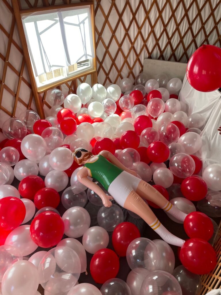 A sad bro'ski looking doll lying sadly in a pile of red and white balloons in a yurt